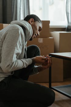 A man assembles a table in his new apartment surrounded by cardboard boxes.